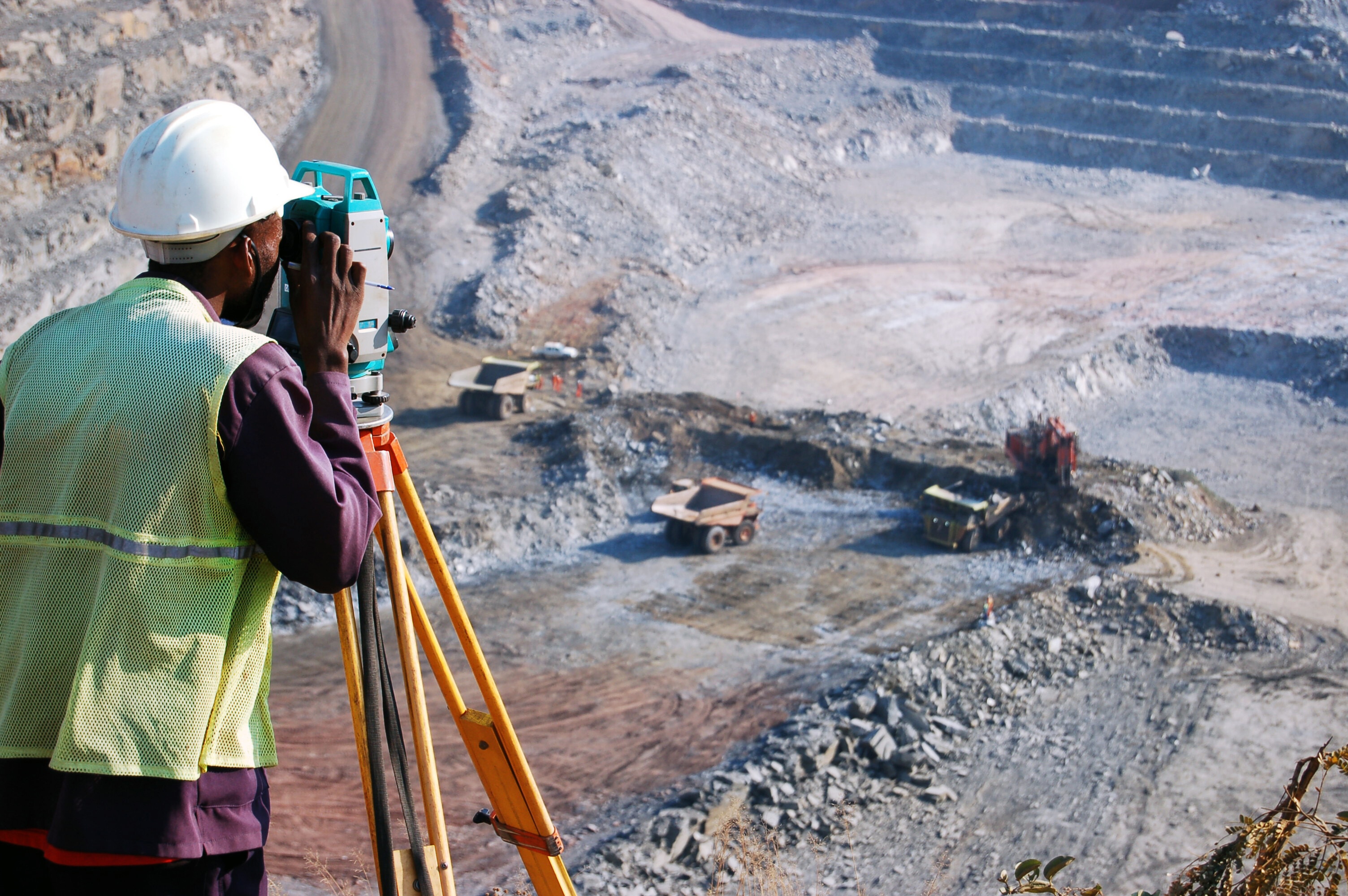 An African man, locally employed as a surveyor at an open-pit copper mine in Zambia, peers through his survey instrument. This work records the daily changes in the open-pit, and help guide mining activities to the engineer's plans. critical minerals