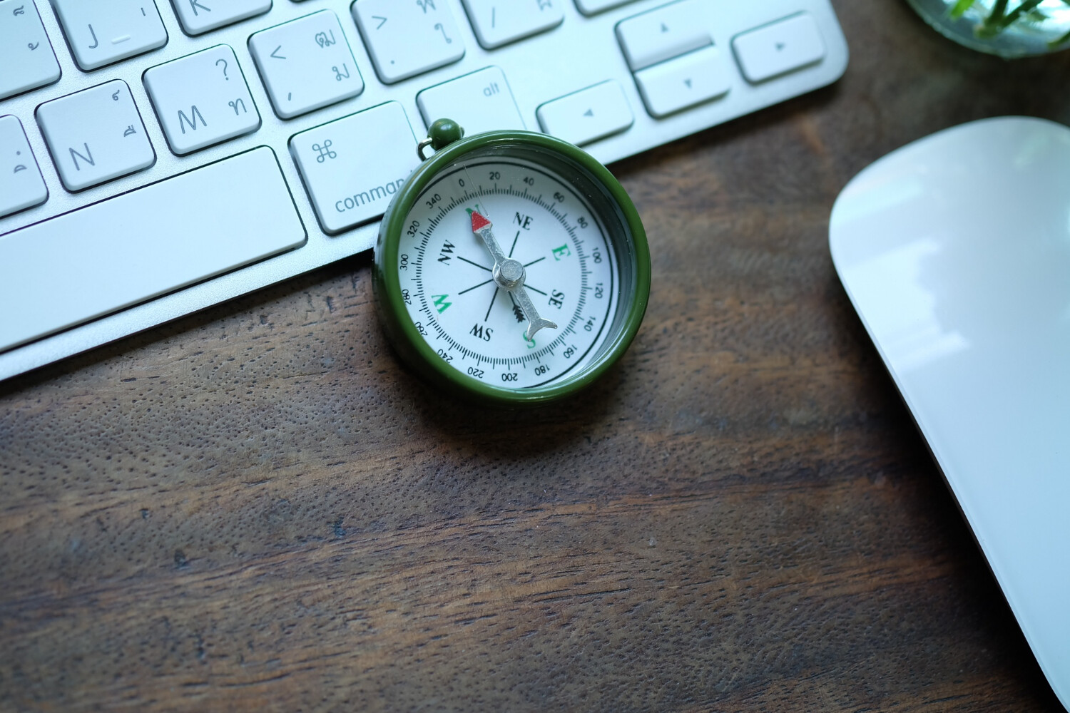A compass placed on a computer keyboard and next to a mouse on a wooden desk.