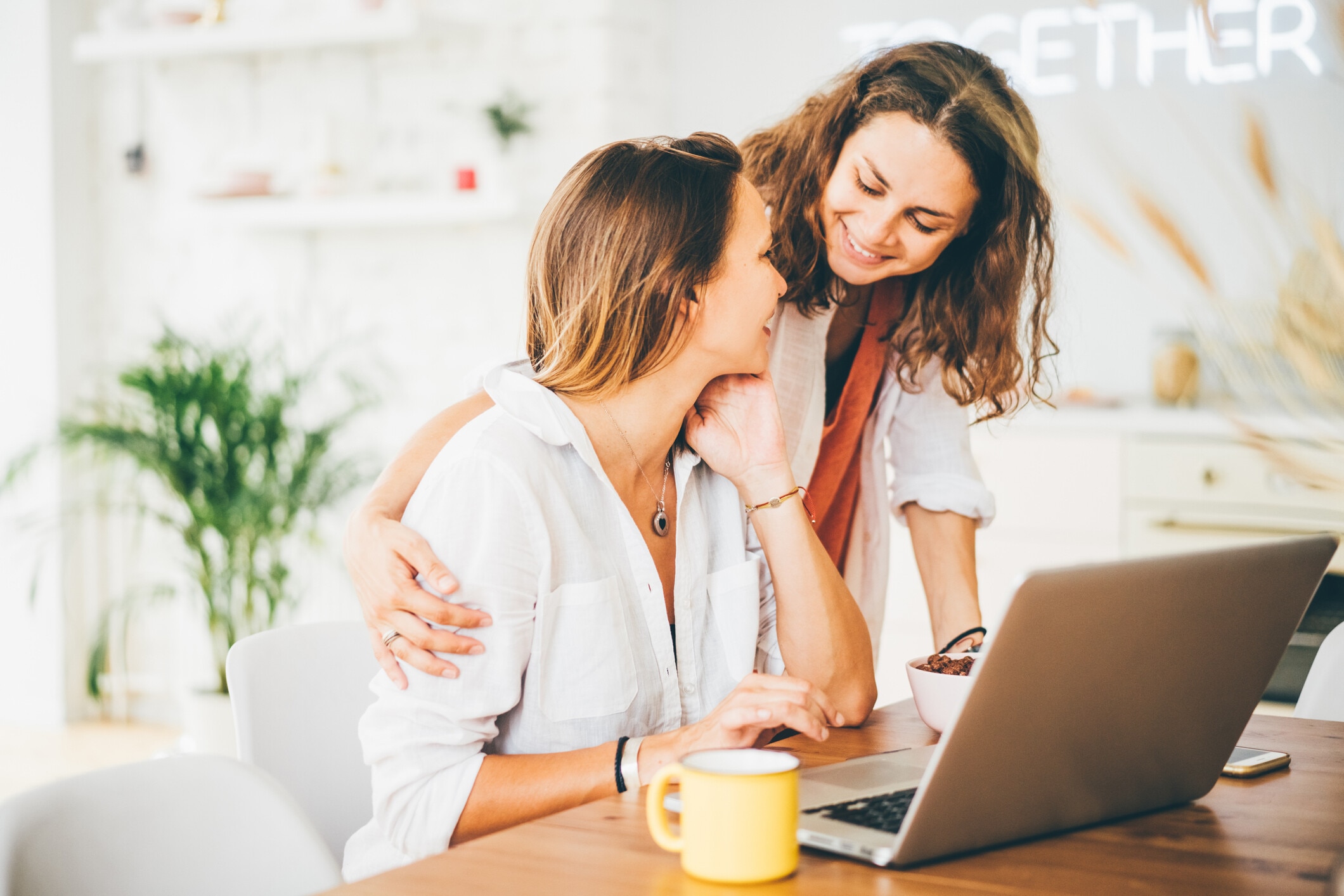 Morning in modern apartment . One girl working on laptop at the kitchen table, her girlfriend comes with breakfast, kisses on the cheek and together watching at display. Concept LGBT couple at home; mental health and wellbeing, family, caring