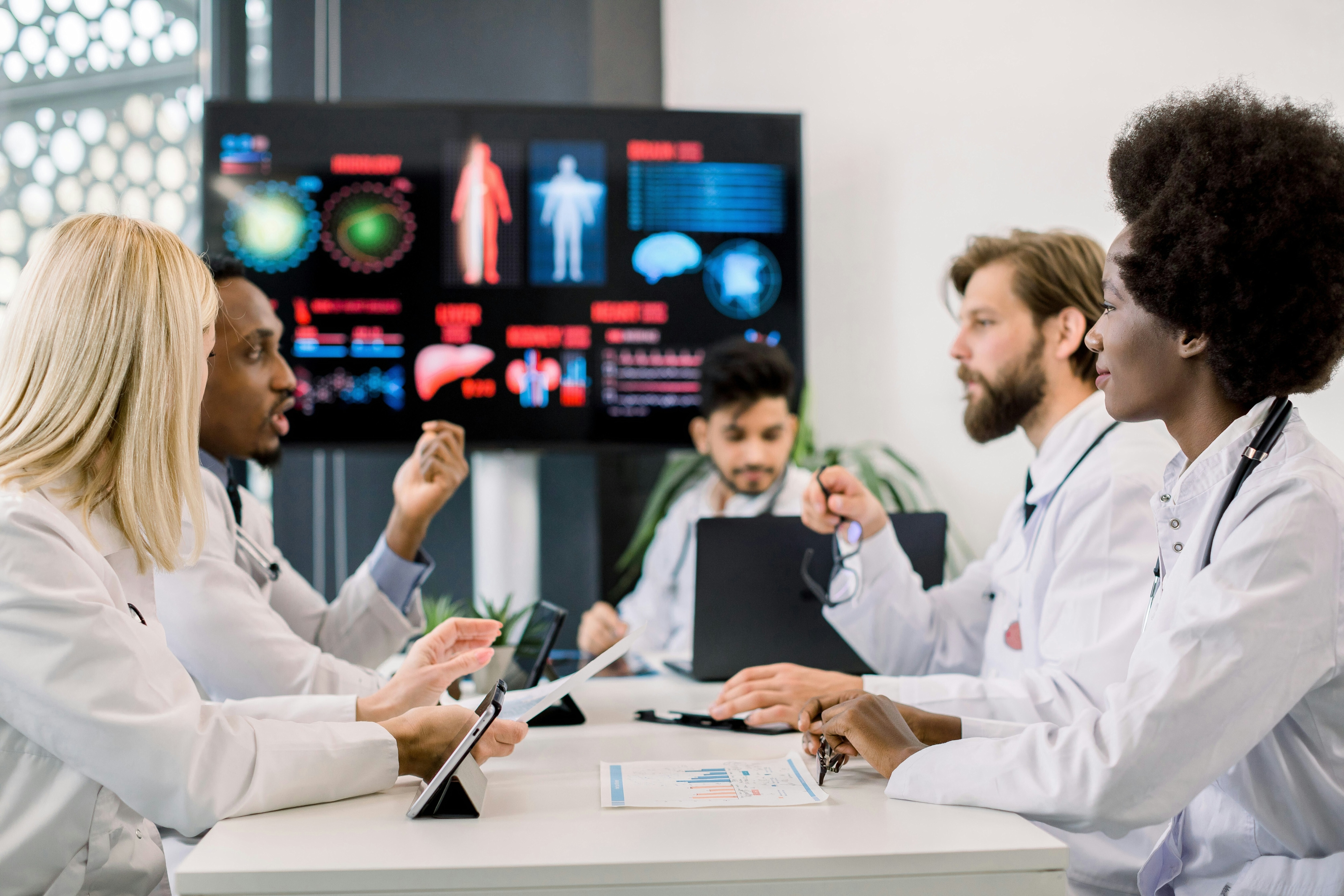 A group of health professionals in white coats and stethoscopes sit around a table with technology/devices: Rebalancing the health data economy requires outcome-based value sharing