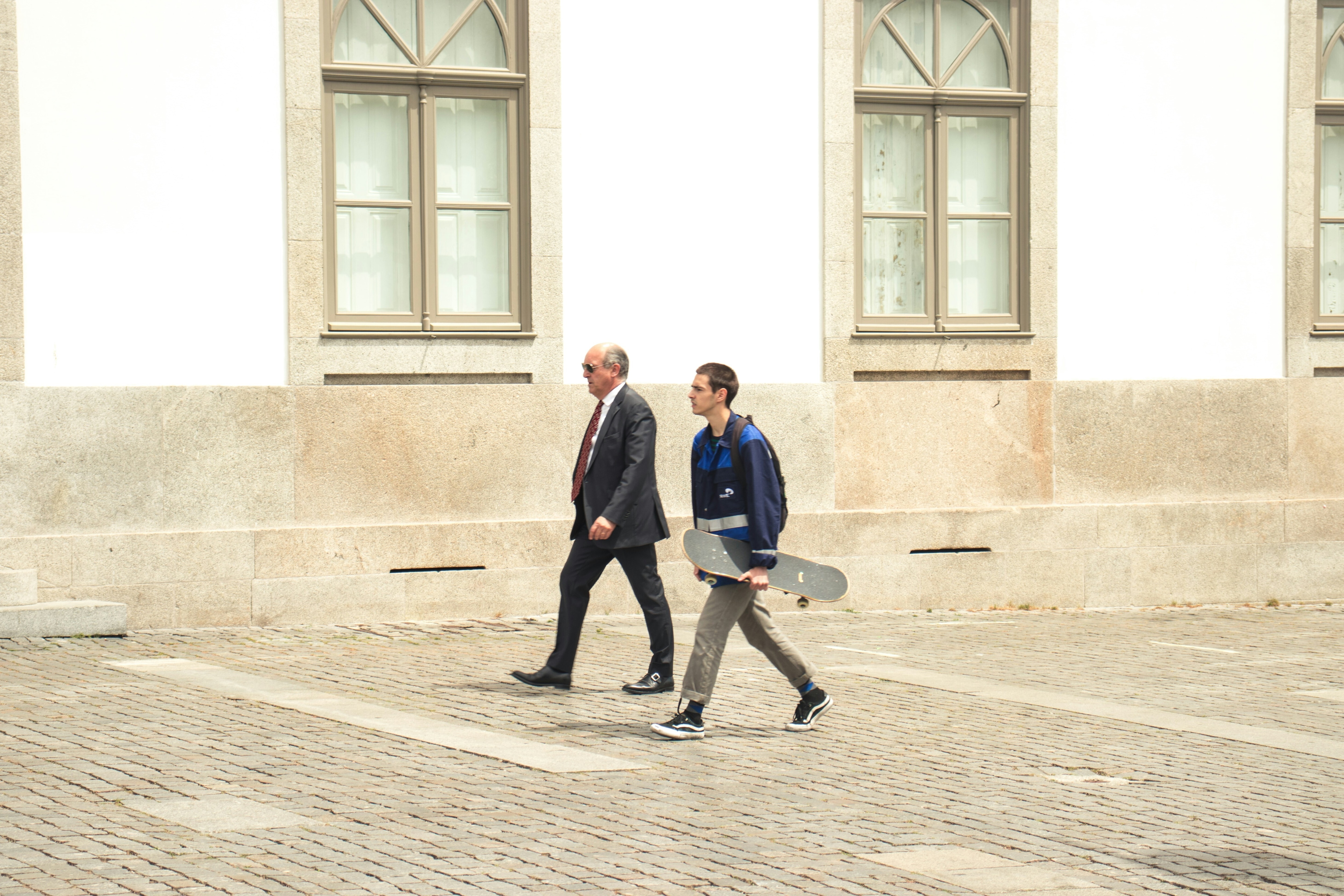 An older man in a suit and a younger man with a skateboard walk along a street; longer lives