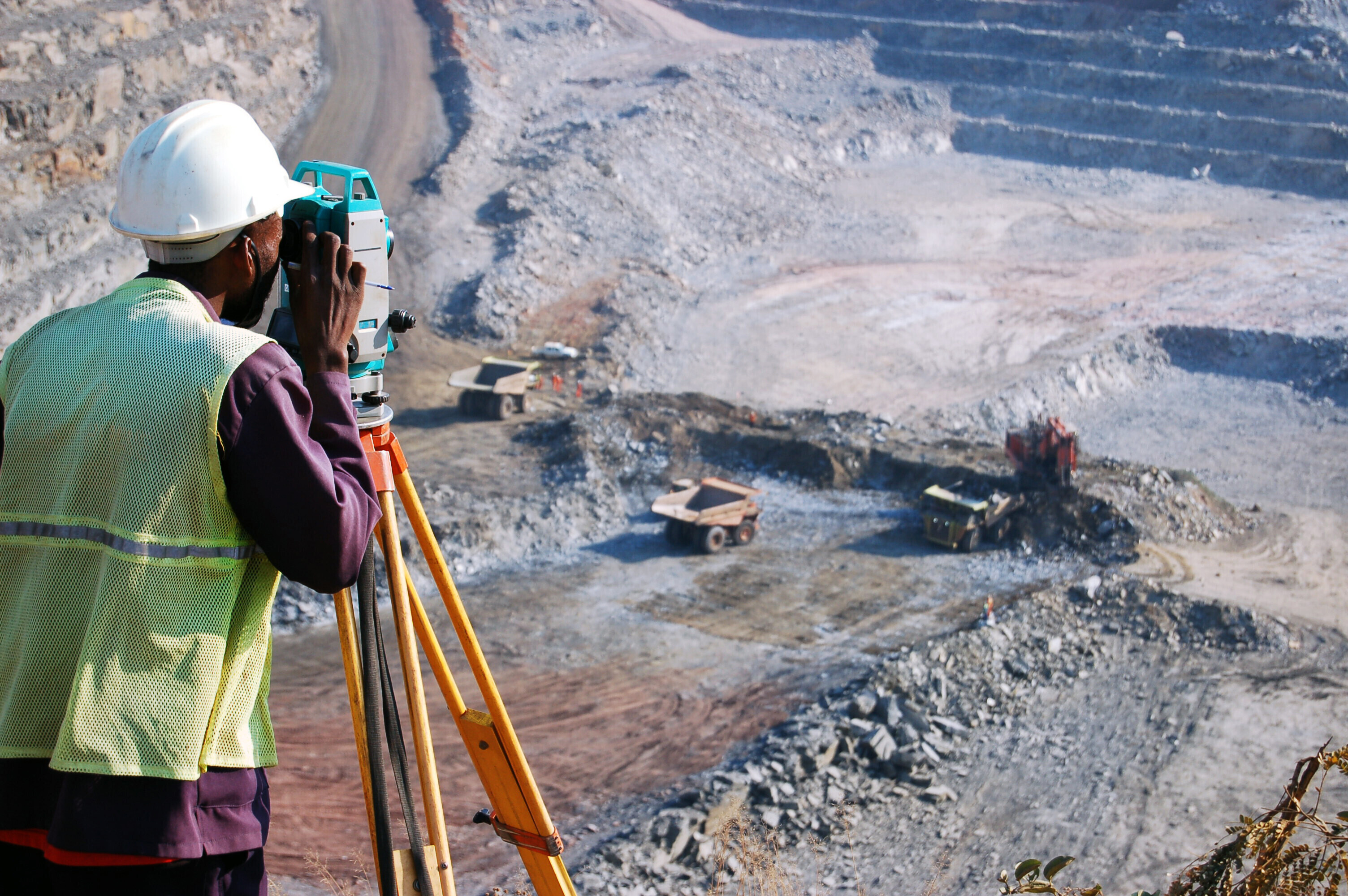 Un hombre africano, empleado localmente como topógrafo en una mina de cobre a cielo abierto en Zambia, observa a través de su instrumento topográfico. Este trabajo registra los cambios diarios en la mina y ayuda a guiar las actividades mineras según los planos del ingeniero.