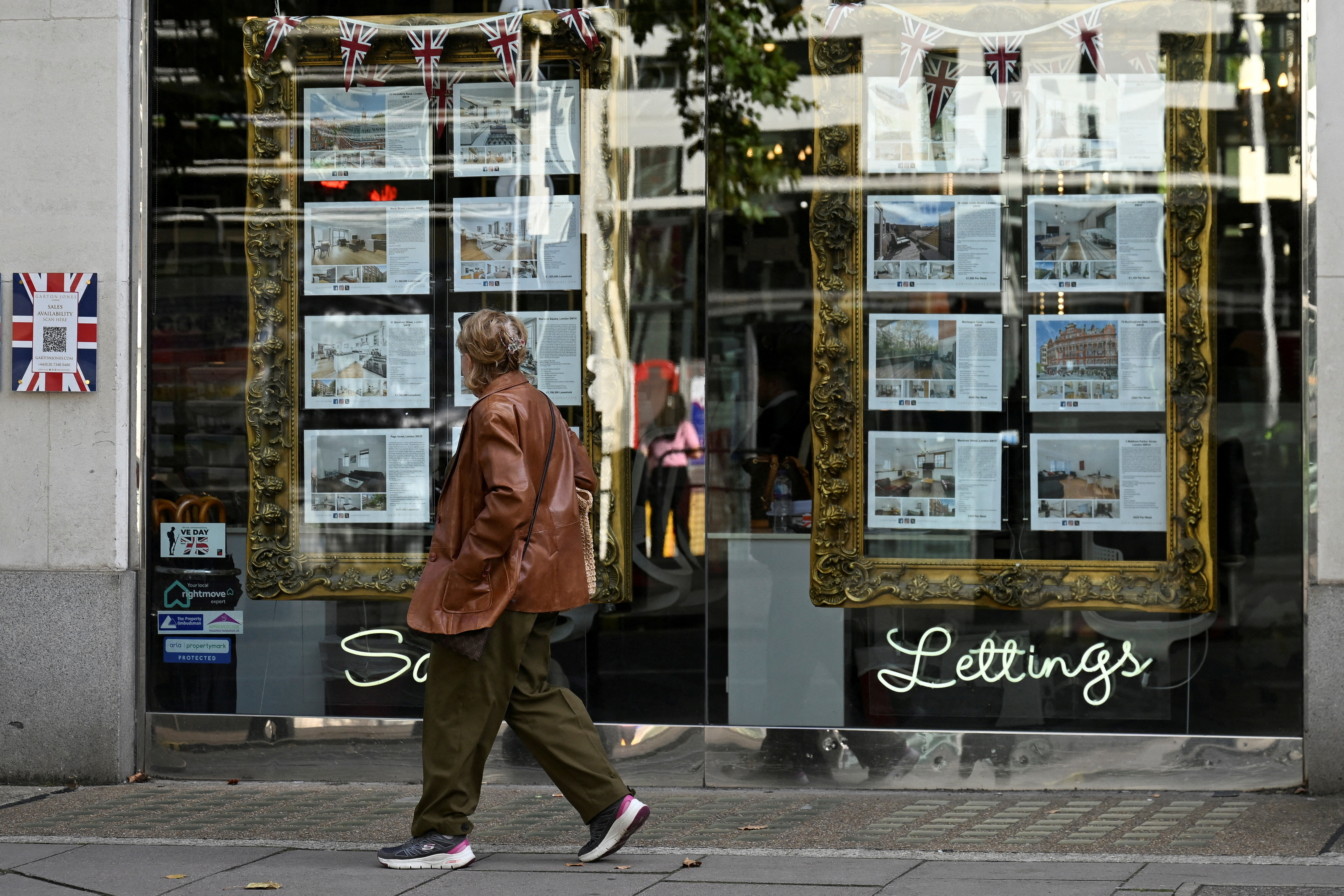 A woman looks at property notices displayed as she walks past a real estate agency in Kensington, in London, Britain, September 17, 2024: We must redesign cities and think beyond affordable housing to address cost of living