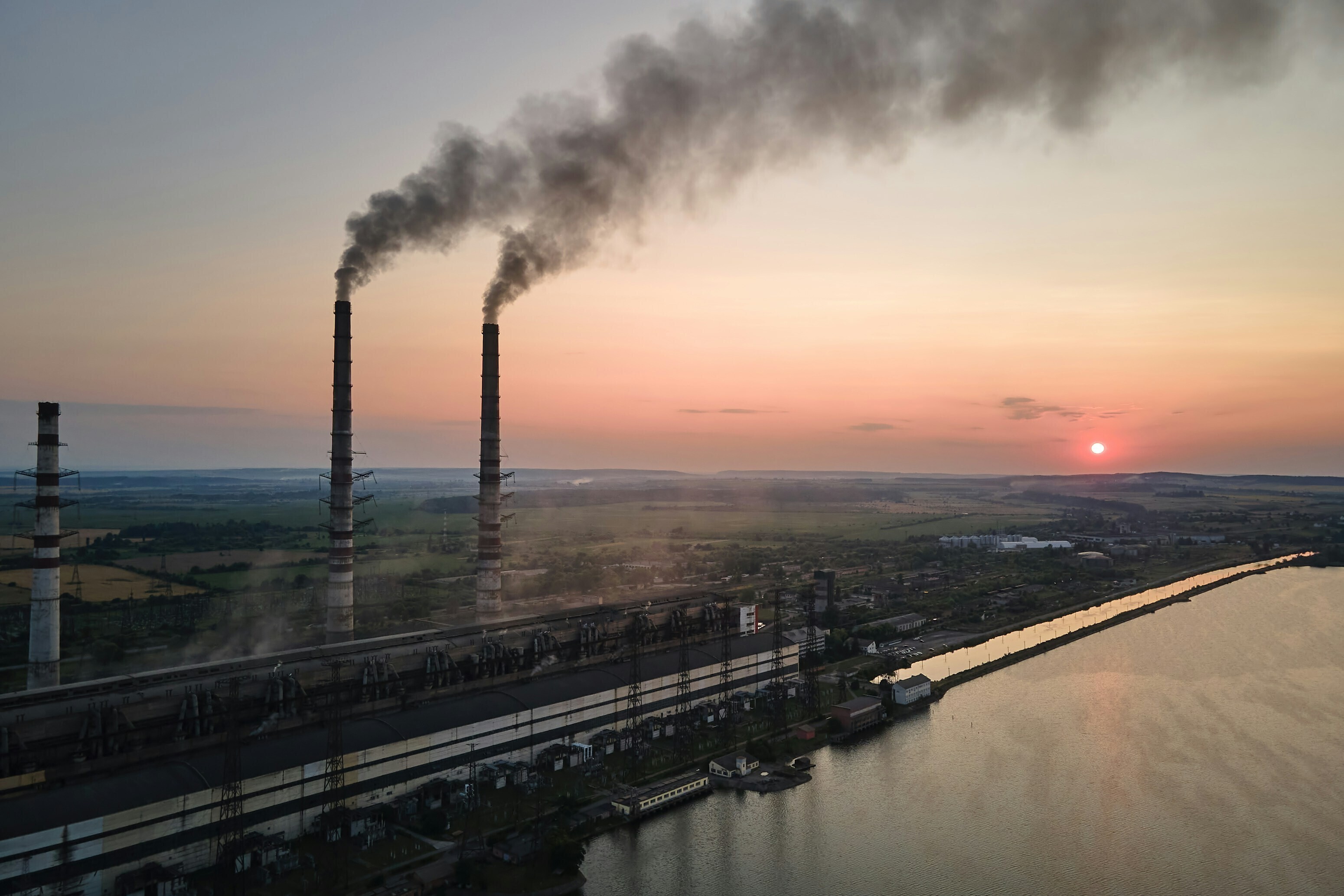 Two industrial chimneys emitting smoke against a pink hued sunset: Higher carbon prices raise capital and deter carbon emissions but carry risks