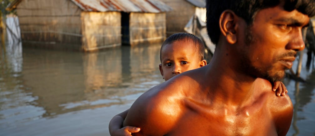 A child reacts to the camera while sitting on his father back as they make their way in a flooded area in Bogra, Bangladesh, August 20, 2017