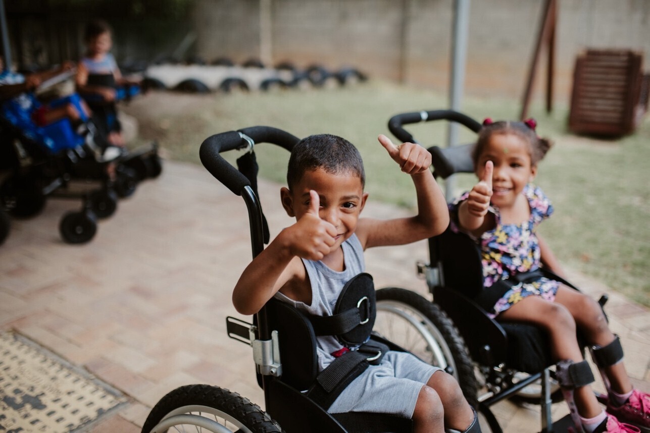 Two children in wheelchairs smile at the camera and give the "thumbs up" sign.