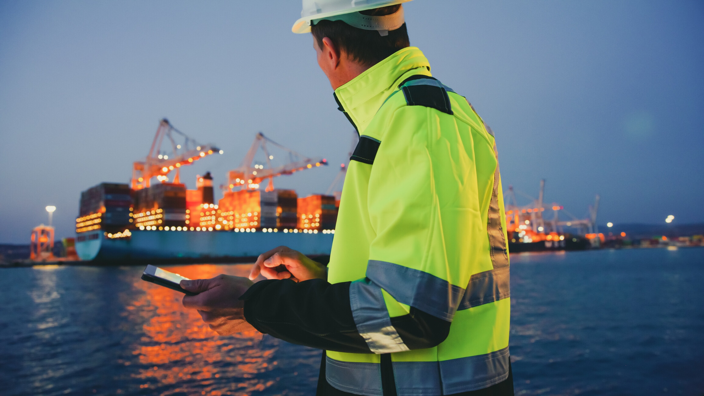 Foreperson in protective gear using digital tablet in front of container terminal port during night; autonomous AI, supply chains