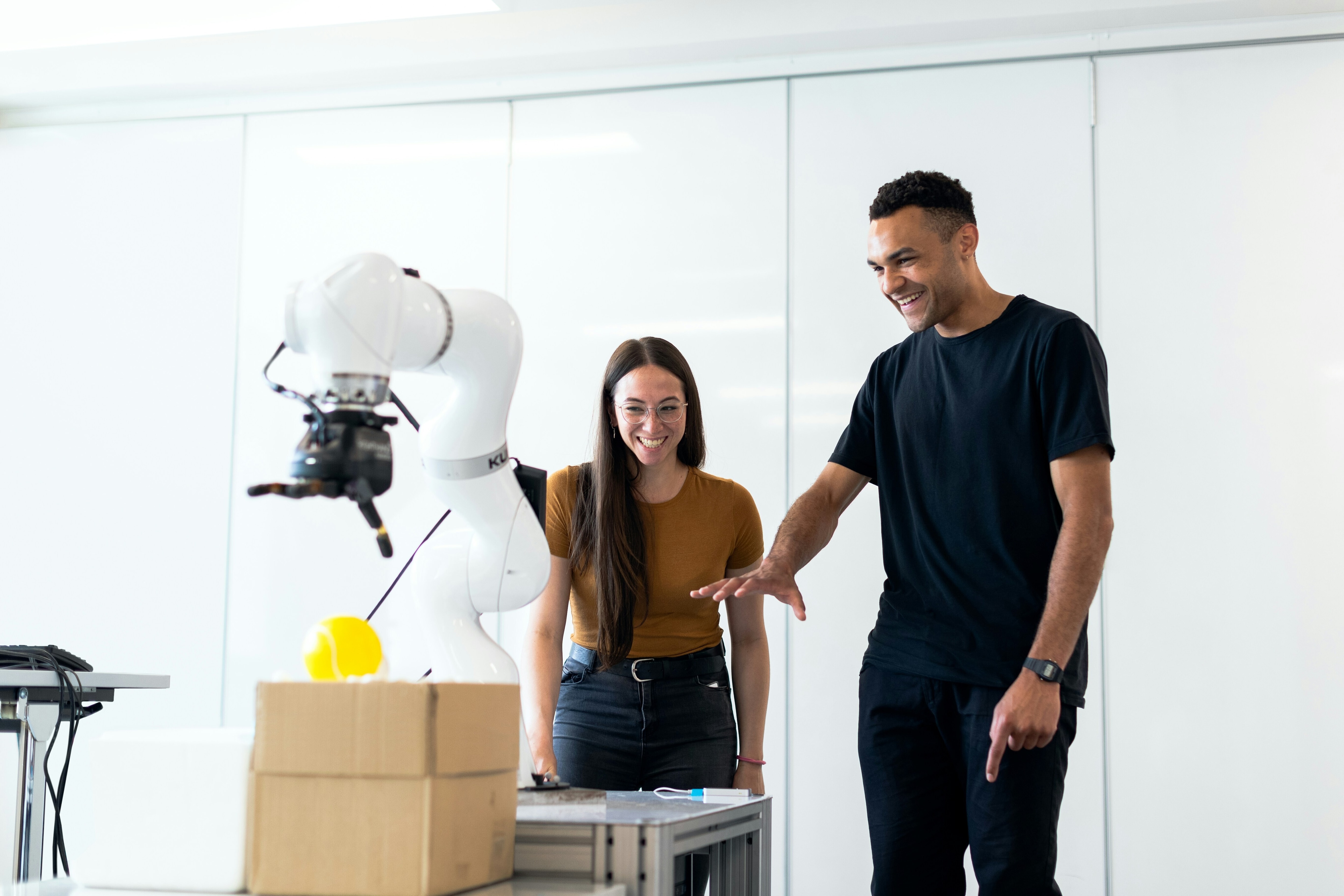 A man and woman look at a robotic arm laughing: The future of work requires human oversight of AI