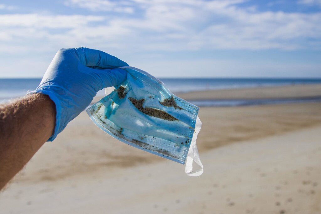A man wearing a blue latex glove holding a mask covered in sand on the beach - and example of medical waste.