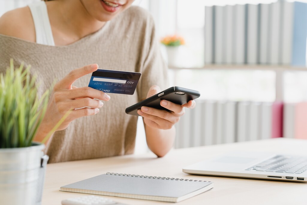 Beautiful Asian woman using smartphone buying online shopping by credit card while wear sweater sitting on desk in living room at home. Lifestyle woman at home concept.