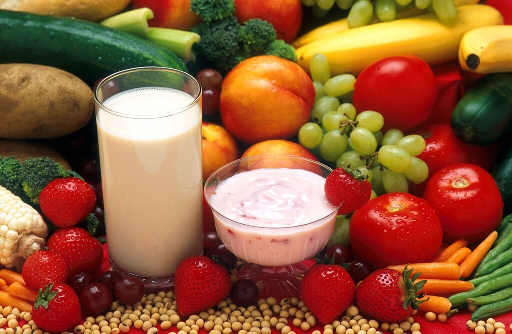 Pictured is a glass of plant-based milk and yoghurt surrounded by colourful fruit and vegetables.