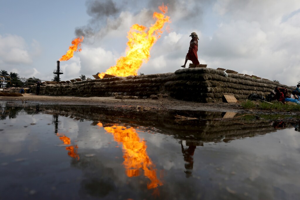 Two gas flaring furnaces and a woman walking on sand barriers are reflected in a pool of oil-smeared water at a flow station in Ughelli, Delta State, Nigeria September 17, 2020. Picture taken September 17, 2020.