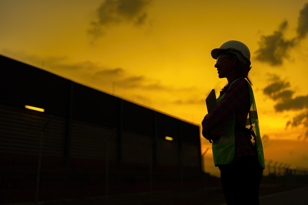 Silhouette of a female engineer smiling. golden sky during sunset standing holding tablet Wear a helmet and a vest as an inspection manager of an industrial factory. about energy and safety