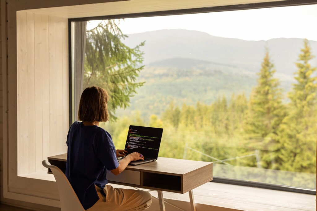 Una persona está sentada en un escritorio escribiendo en una computadora portátil. La gran ventana muestra una vista de montañas y árboles a lo lejos. La escena es luminosa y acogedora.