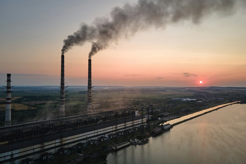 Two industrial chimneys emitting smoke against a pink hued sunset: Higher carbon prices raise capital and deter carbon emissions but carry risks