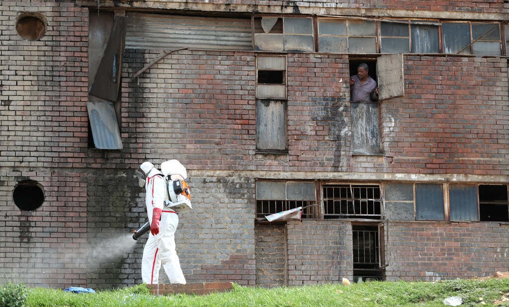 A resident looks out of the window as a worker wearing protective gear sprays disinfectant to sanitise parts of Madala Men's Hostel during a 21-day nationwide lockdown to try to contain the coronavirus disease (COVID-19) outbreak, in Alexandra, South Africa April 1, 2020. REUTERS/Siphiwe Sibeko - RC2QVF9615M1