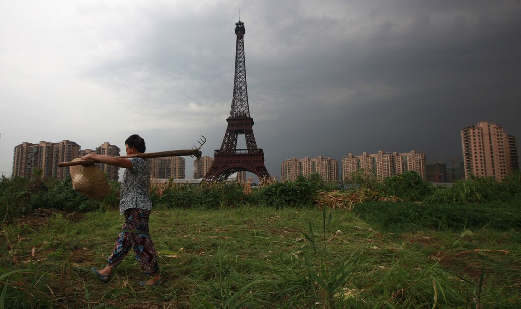 A farmer carrying a rake walks down a dirt road past a replica of the Eiffel Tower at the Tianducheng development in Hangzhou, Zhejiang Province August 1, 2013. Tianducheng, developed by Zhejiang Guangsha Co. Ltd., started construction in 2007 and was known as a knockoff of Paris with a scaled replica of the Eiffel Tower standing at 108 metres (354 ft) and Parisian houses. Although designed to accommodate at least 10 thousand people, Tianducheng remains sparsely populated and is now considered as a "ghost town", according to local media. REUTERS/Aly Song (CHINA - Tags: SOCIETY BUSINESS REAL ESTATE AGRICULTURE TPX IMAGES OF THE DAY) - RTX127WU