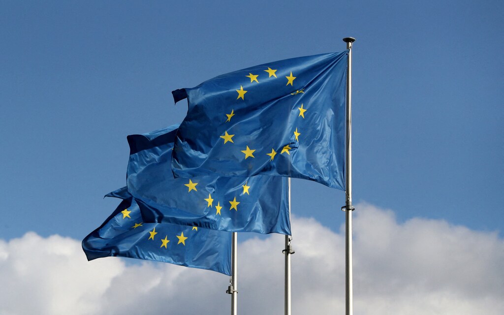 Three European Union flags are seen against the backdrop of a blue, slightly cloudy sky.