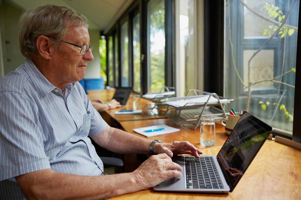 A senior gentleman in a striped blue shirt working at a desk on a laptop with coworkers in the background: Encouraging an age-inclusive workforce could be essential to unlocking the longevity economy