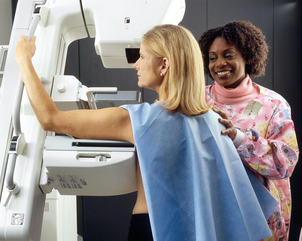 Smiling health professional assists woman in scrubs having a mammogram: Women’s health is still not cemented in national policy decisions.
