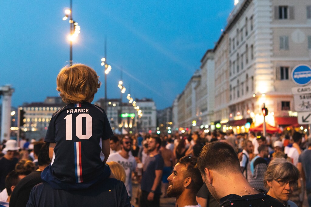 a boy with a France sports shirts sits on an adults shoulders looking out to a crowd