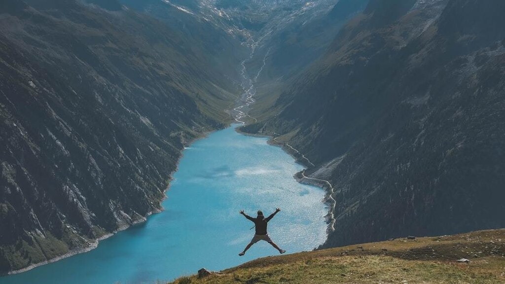 Man doing a star-jump in front of large body of water and mountains.