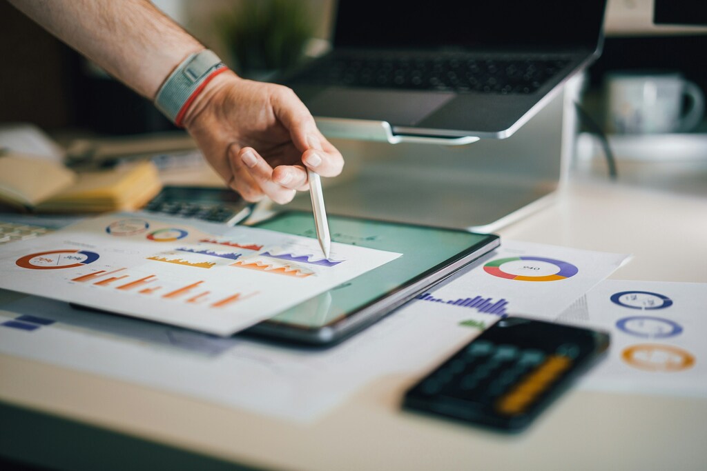 A person analyzing graphs on paper with tablet, calculator and laptop in shot: Implementing impact-linked compensation in impact investing fosters greater accountability among fund managers.
