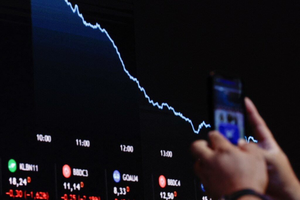 A person holds a mobile phone near an electronic board displaying information on recent fluctuations of market indices at the B3 Stock Exchange in Sao Paulo, Brazil April 4, 2025. REUTERS/Amanda Perobelli