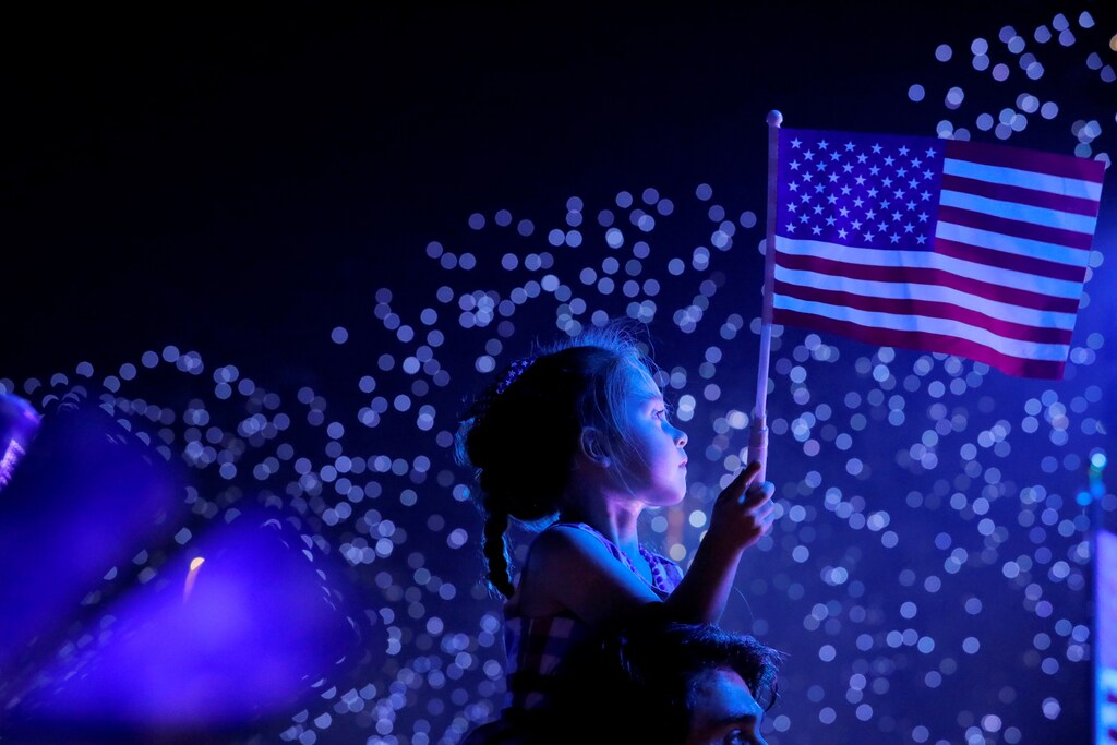 A child watches a Fourth of July fireworks in New York City, while holding an American flag.