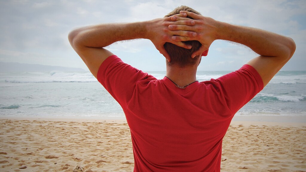 Hombre con camiseta roja y las manos detrás de la cabeza mirando al mar en la playa: El fitness mental implica un enfoque coherente de la salud mental.