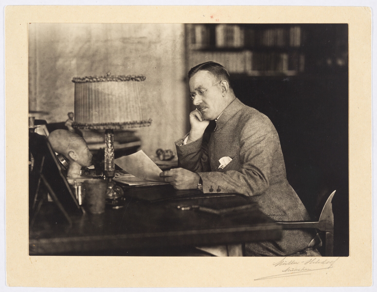 Thomas Mann, Brustbild, Halbprofil nach links, mit Brille, Kopf auf die rechte Hand gestützt, Manuskript in der linken Hand haltend.