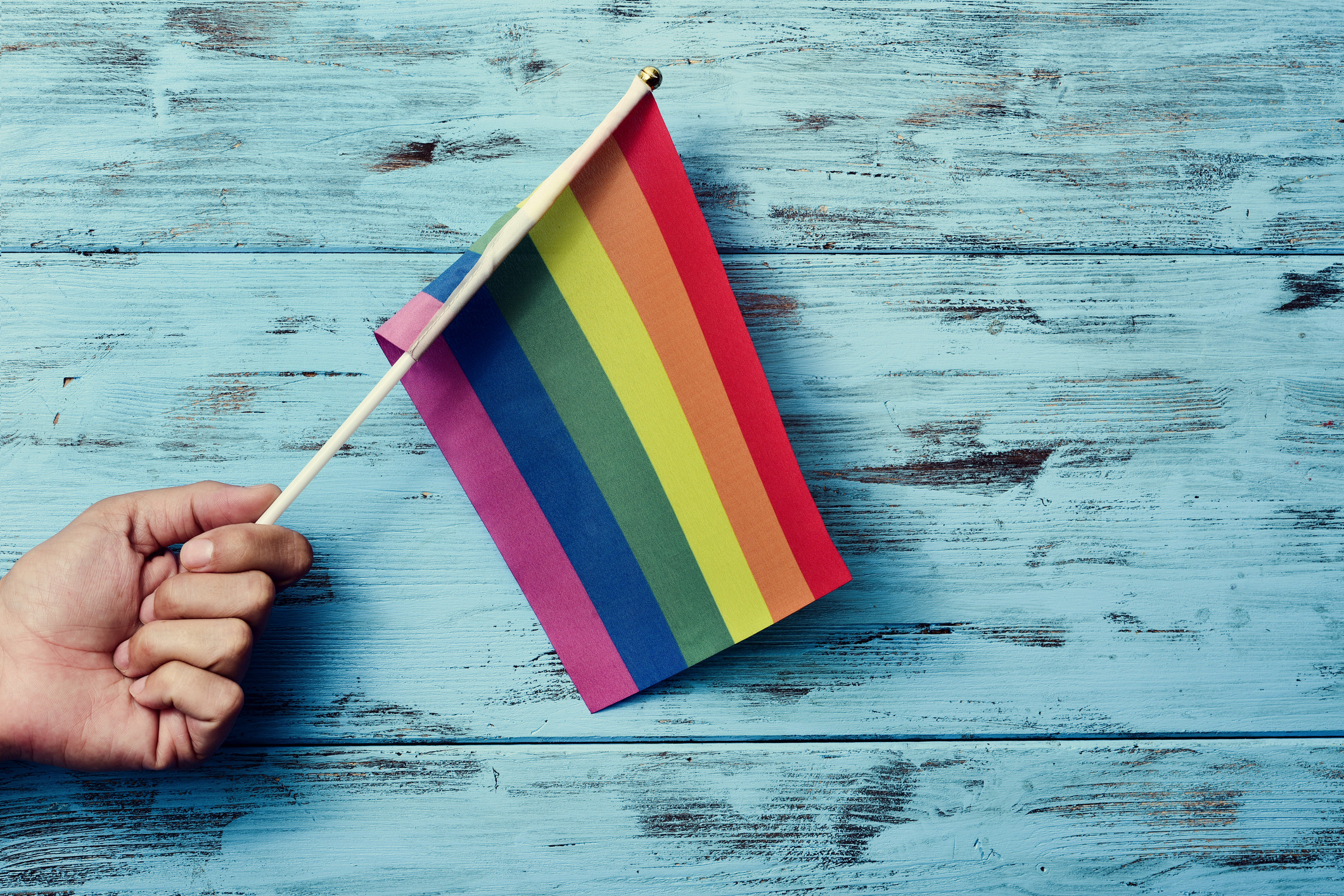 El primer plano de un joven caucásico ondeando una pequeña bandera del arco iris frente a un fondo rústico de madera azul      